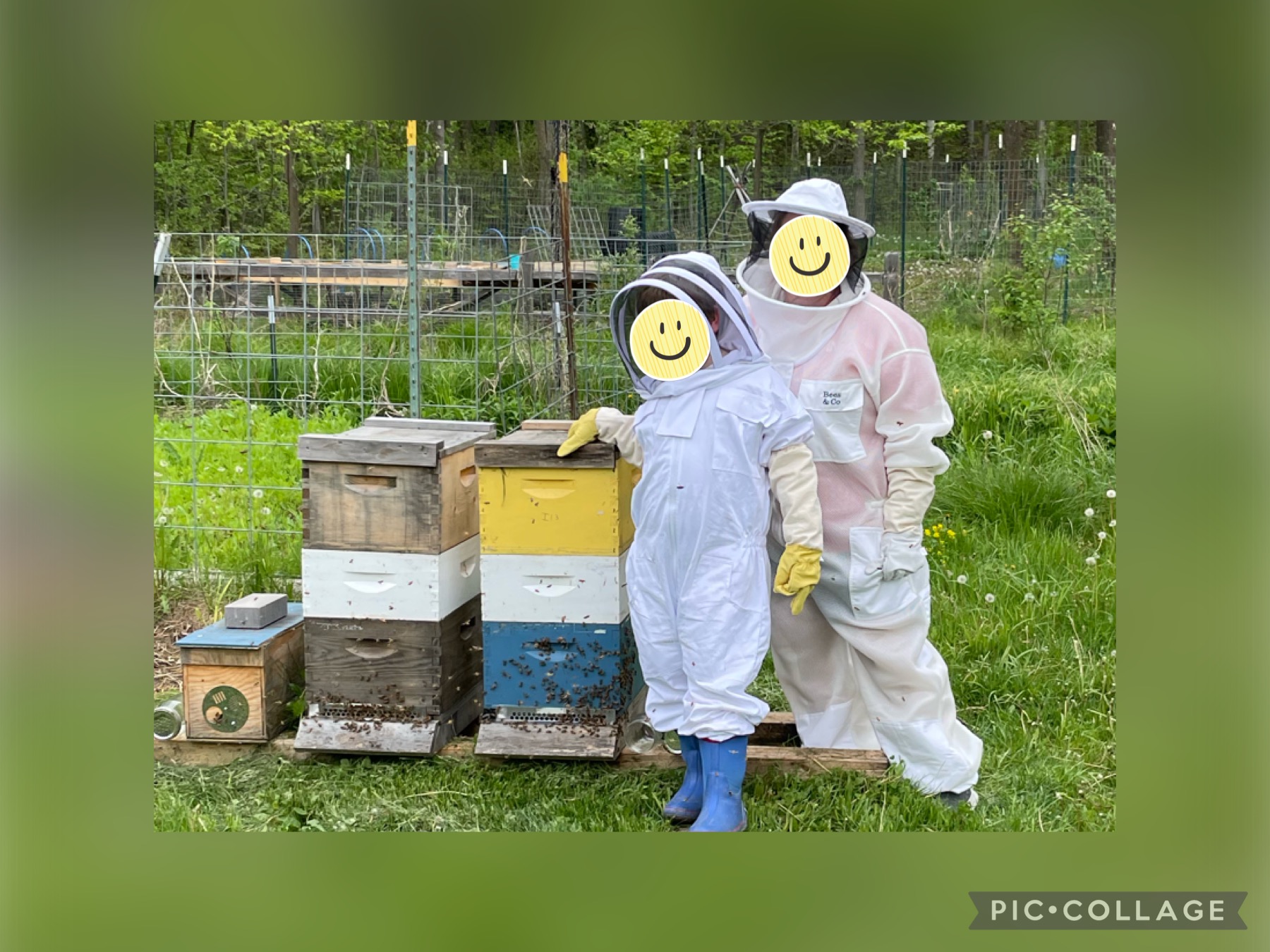 Image shows a mom and her son in bee suits standing next to three honeybee hives.  Smiley face stickers cover their faces to protect their privacy. You can't see the smiles on their faces but their stance shows they are comfortable being close to the hives